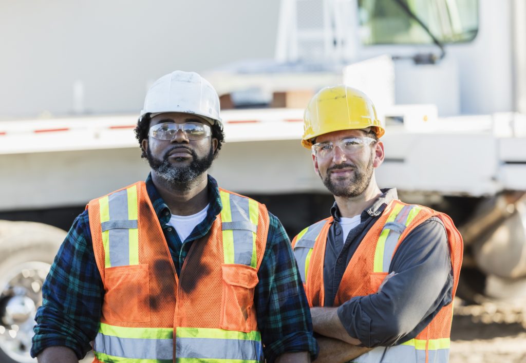 Two multi-ethnic mid-adult men in their 30s, construction workers at a job site. They are wearing hardhats, reflective vests and safety glasses, standing in front of a crane.